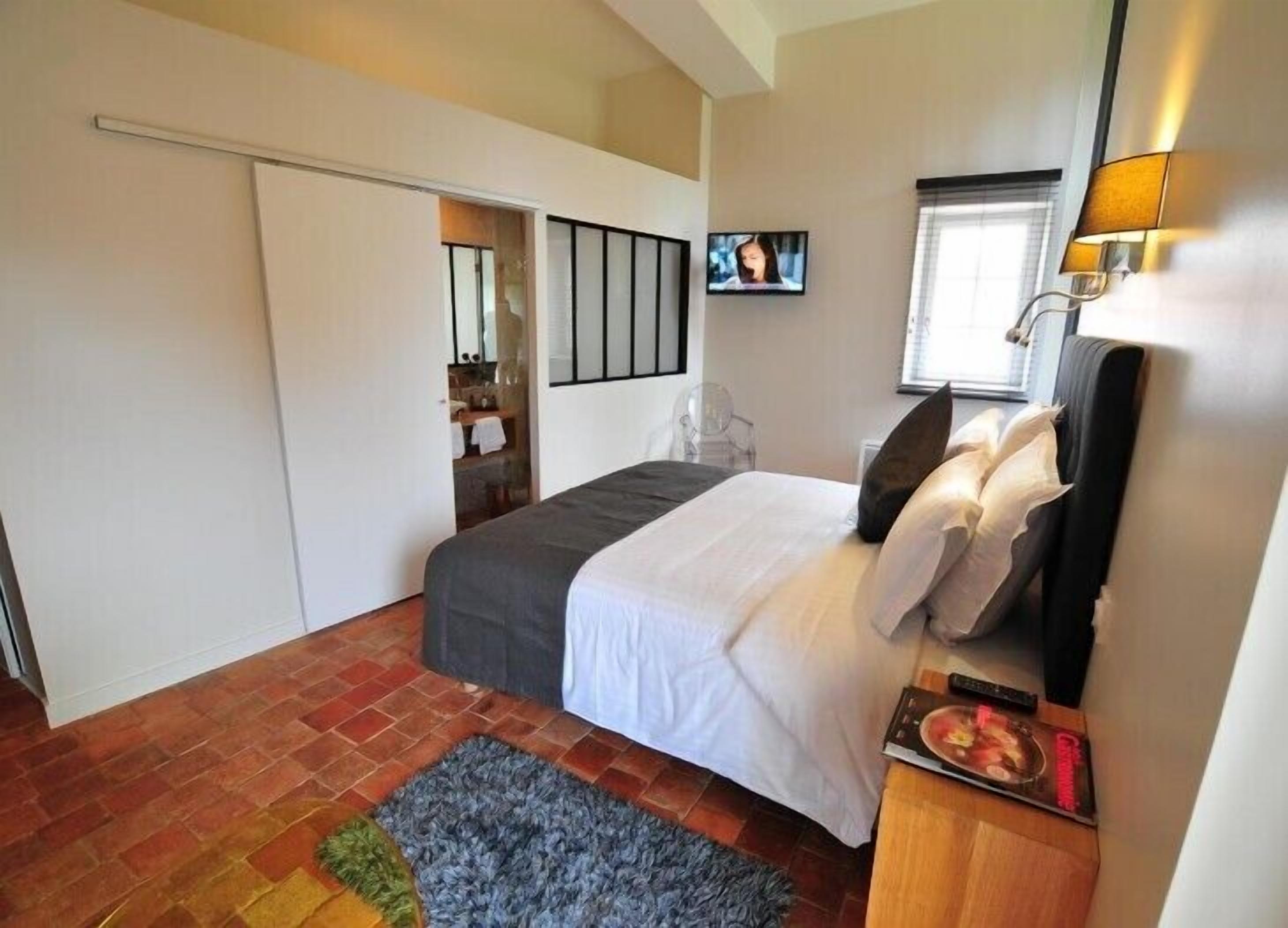 Une chambre à coucher comprenant un lit, un téléviseur, une fenêtre avec des stores, une table de chevet et une porte donnant sur une salle de bain.