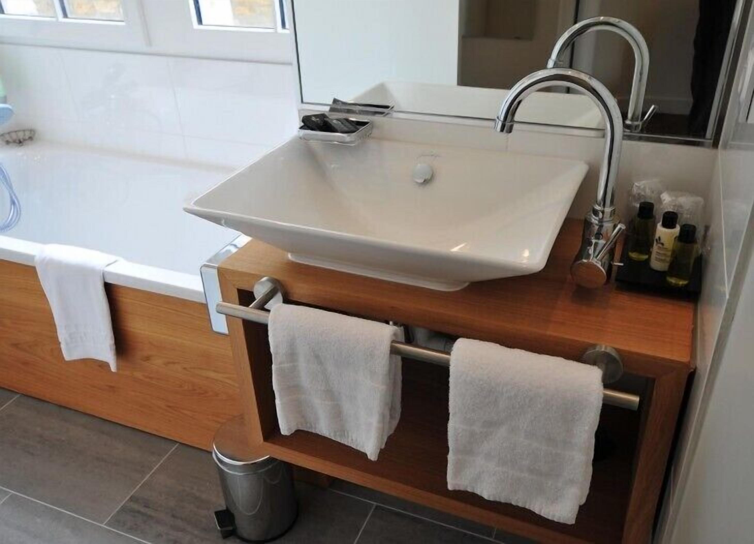 A modern bathroom with a large white sink, a wooden vanity, two towels, a small trash can, and a mirror above the sink.