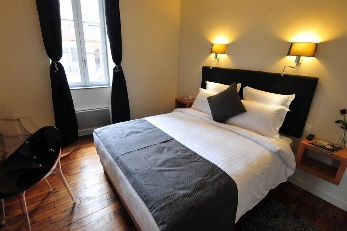 A neatly made bed with a black headboard, two bedside lamps, a small bedside table with a book, and a chair.