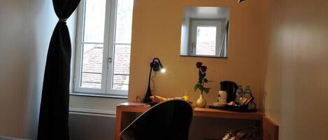 Een hotelkamer met een bed, een bureau met een stoel, een bureaulamp, een vaas met een bloem, een koffiezetapparaat en een handtas op een nachtkastje.