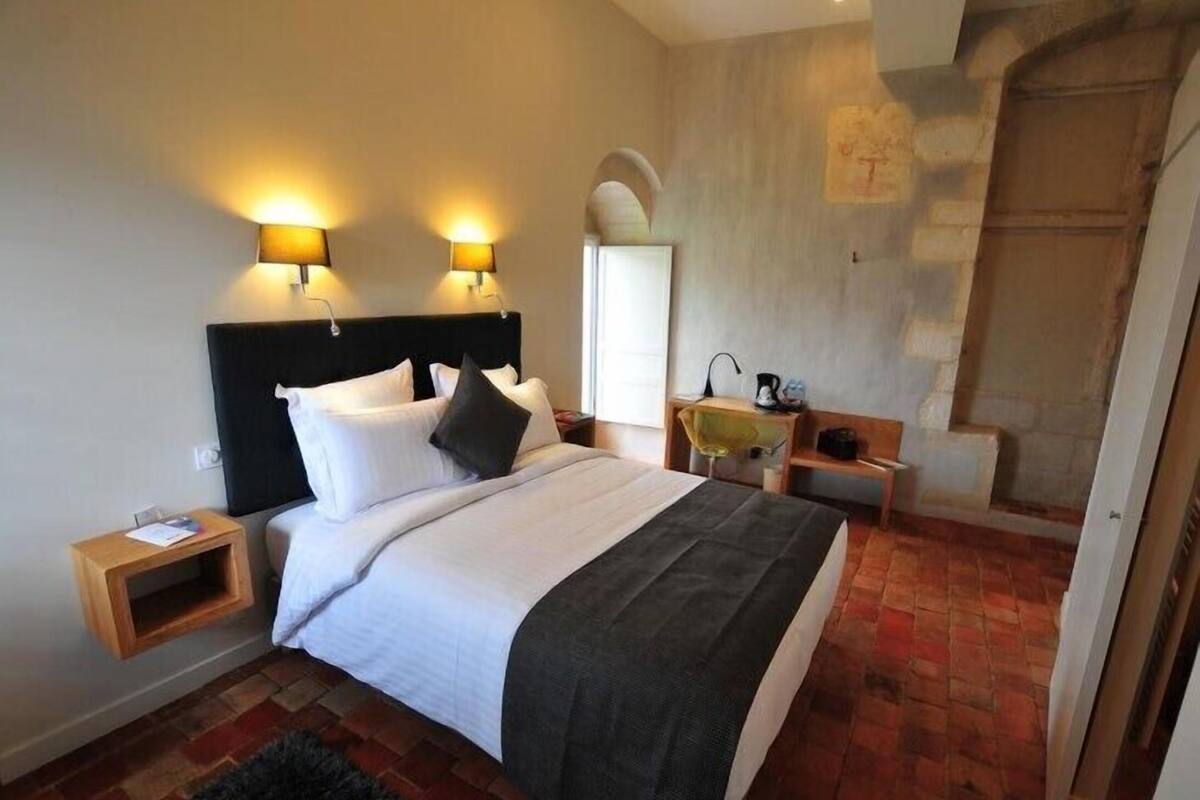 A bedroom with a large bed, two bedside tables, a nightstand, a wooden bench, a stone wall, and a staircase.