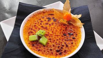 A bowl of crème brûlée with a caramelized top, garnished with kiwi slices and a cherry, served on a white plate with a black napkin.