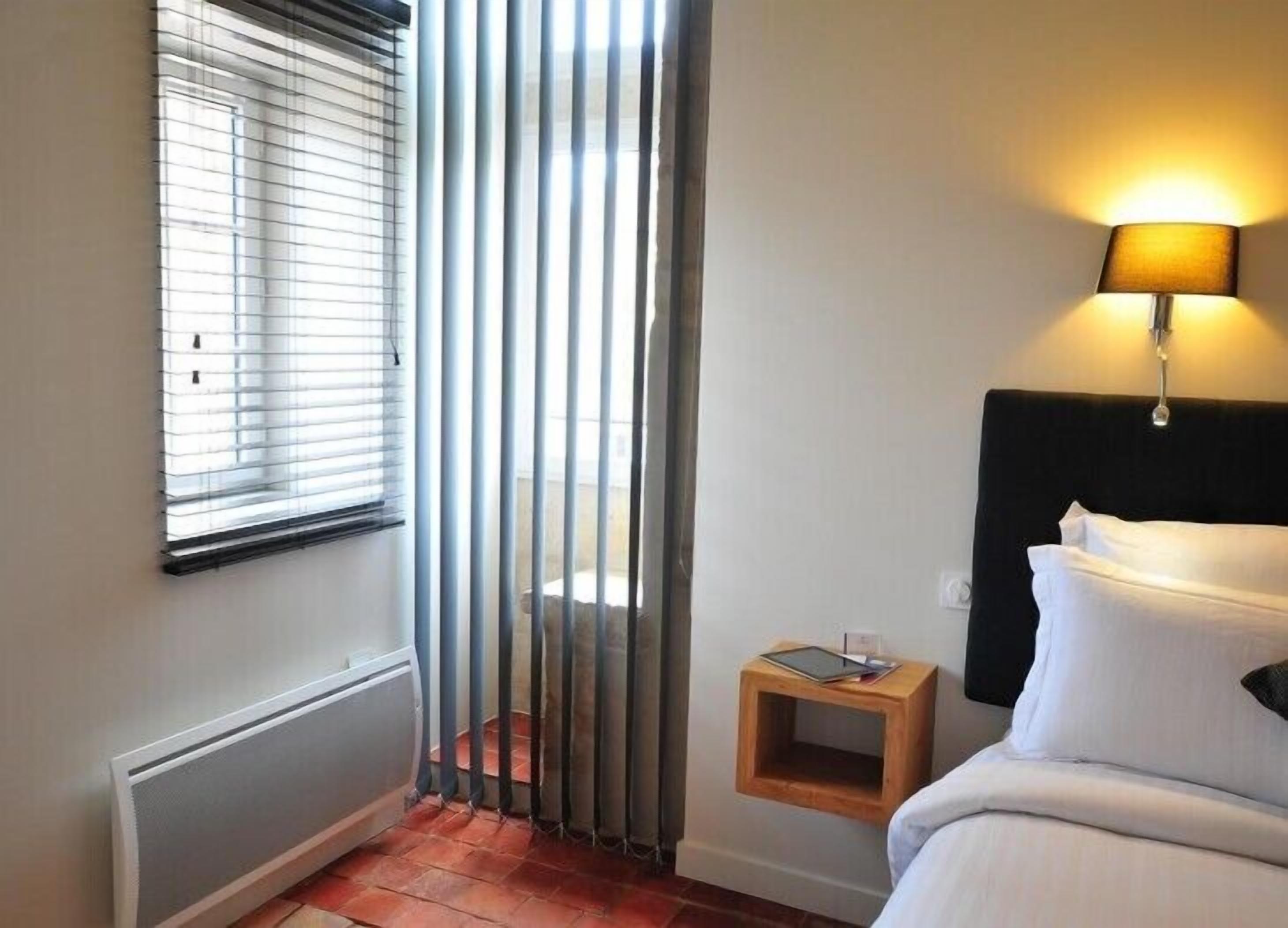 A hotel room with a bed, a bedside table with a lamp, a window with blinds, and a radiator.