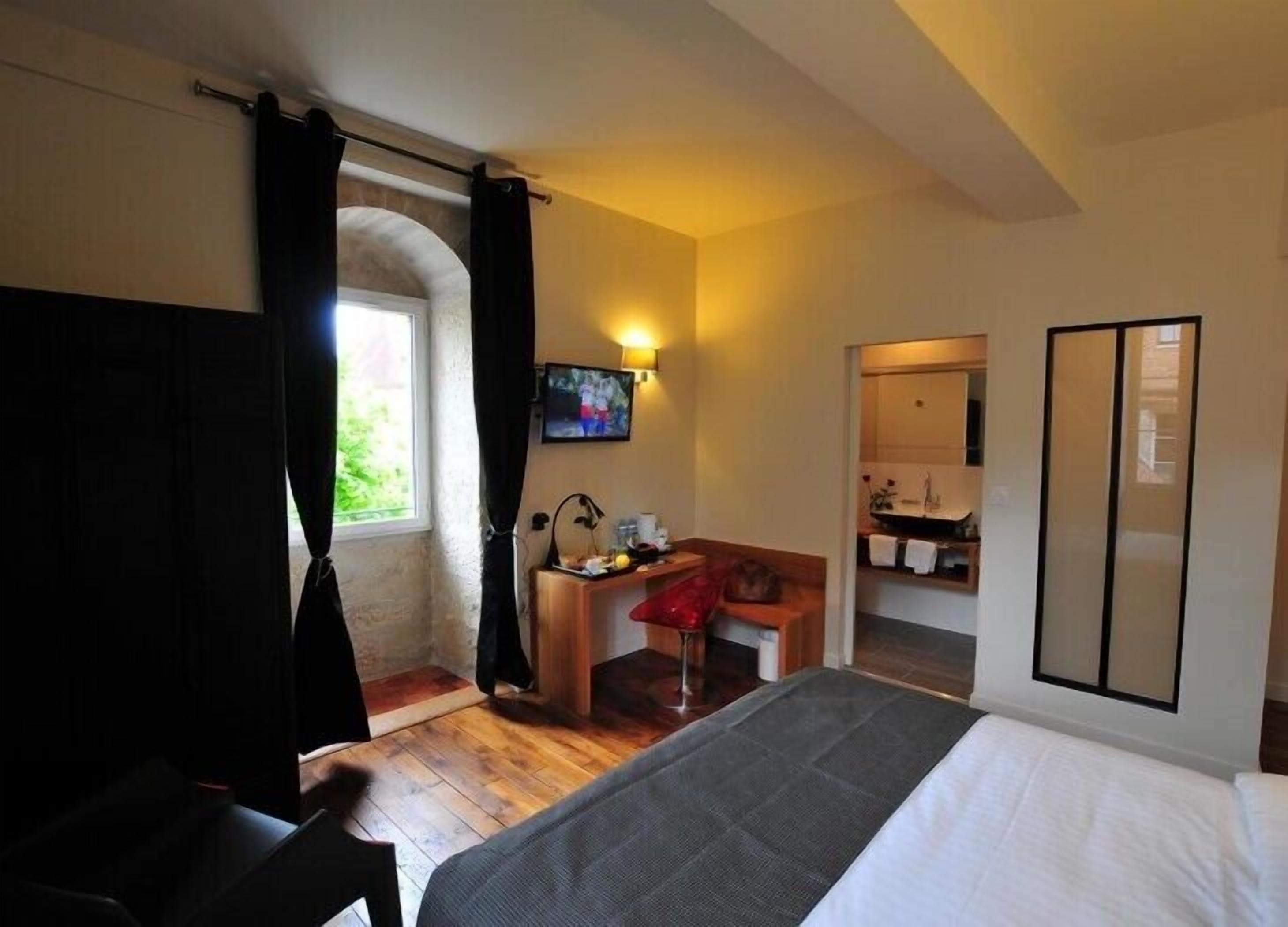 A hotel room with a bed, a desk, a chair, a TV, and a bathroom visible through an archway.