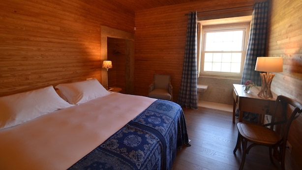 Een kamer met houten vloeren, een bed, een stoel, een tafel en een raam met gordijnen.