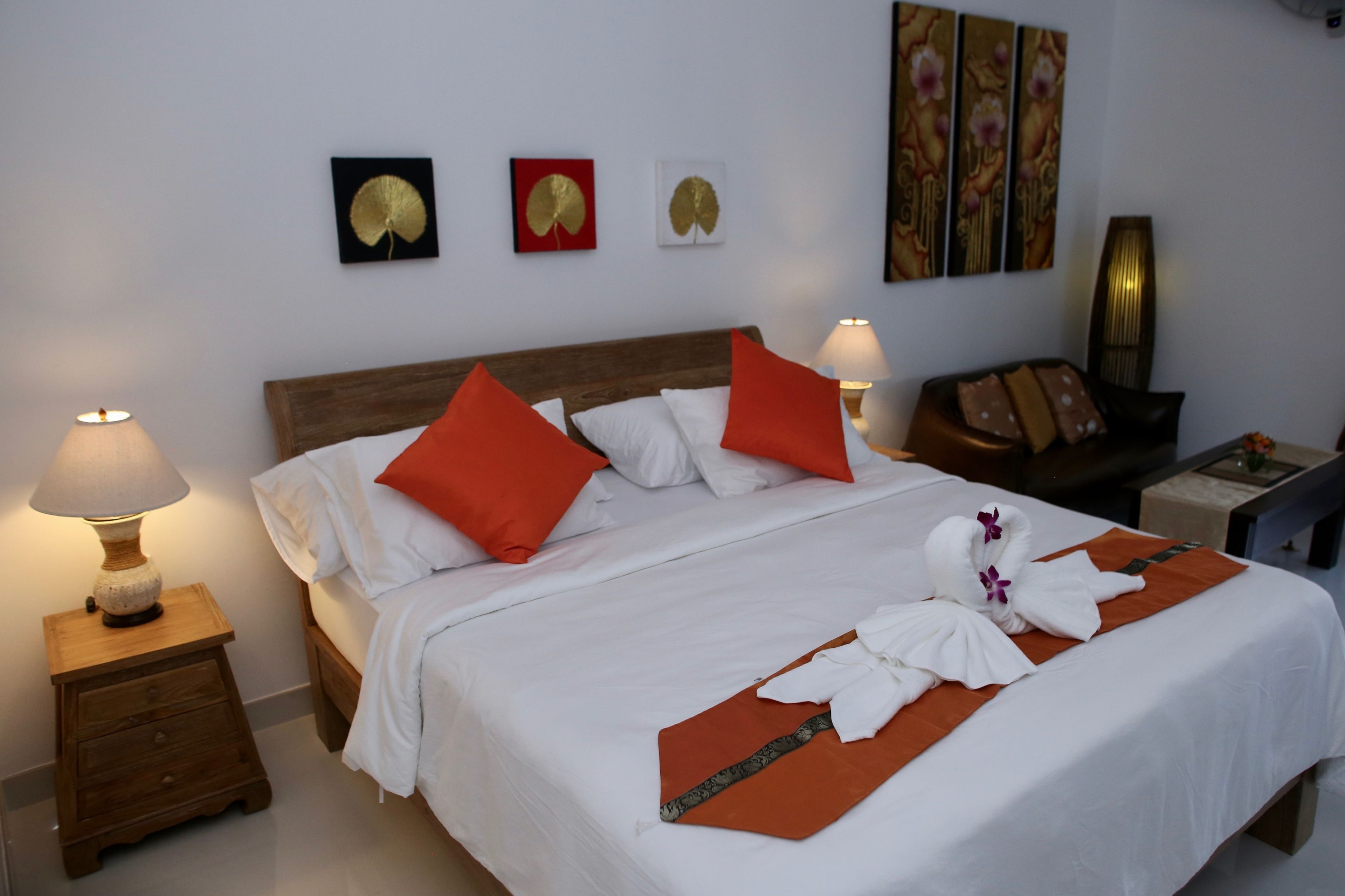 A neatly made bed with white and orange pillows, a wooden headboard, and a decorative towel arrangement. There are wall art pieces and a lamp on the side table.