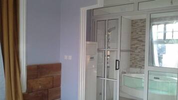 Ein Zimmer mit einem Bett, einem Holz-Kopfteil, einer Glastür zu einem Badezimmer mit gefliestem Duschbereich und einem Fenster mit Vorhängen.