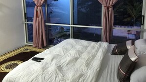 A bedroom with a large window, a bed with white bedding, and a remote control on the bed. - Volta Escape Resort (Atimpoku)