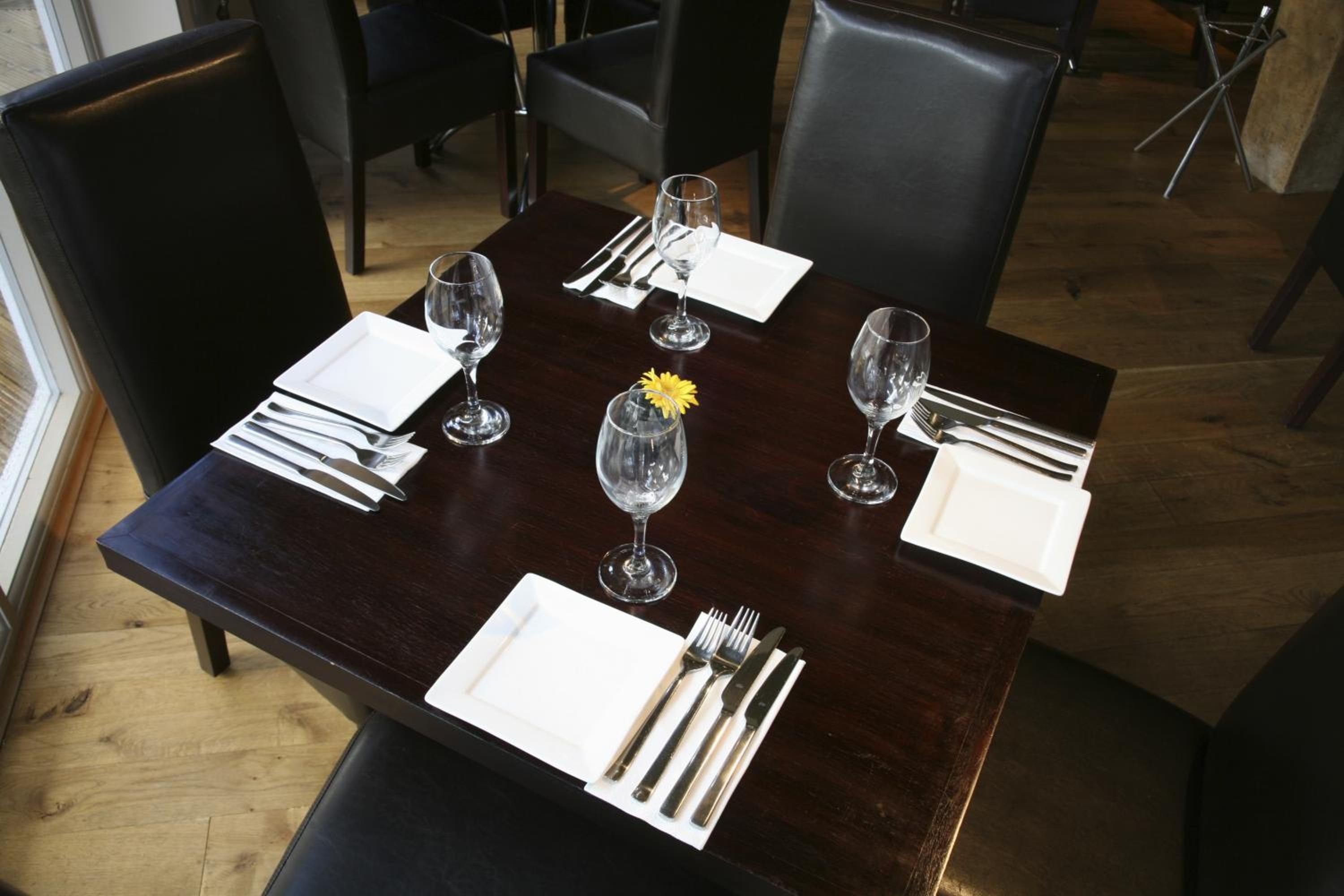 A dining table set with white plates, cutlery, and clear wine glasses, with a single yellow flower centerpiece.
