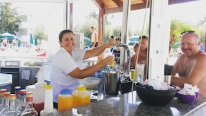 A bar area with a bartender preparing drinks, a counter with bottles and glasses, and a view of a pool area with people in the background. - Cyprotel Faliraki - All Inclusive (Rhodes)