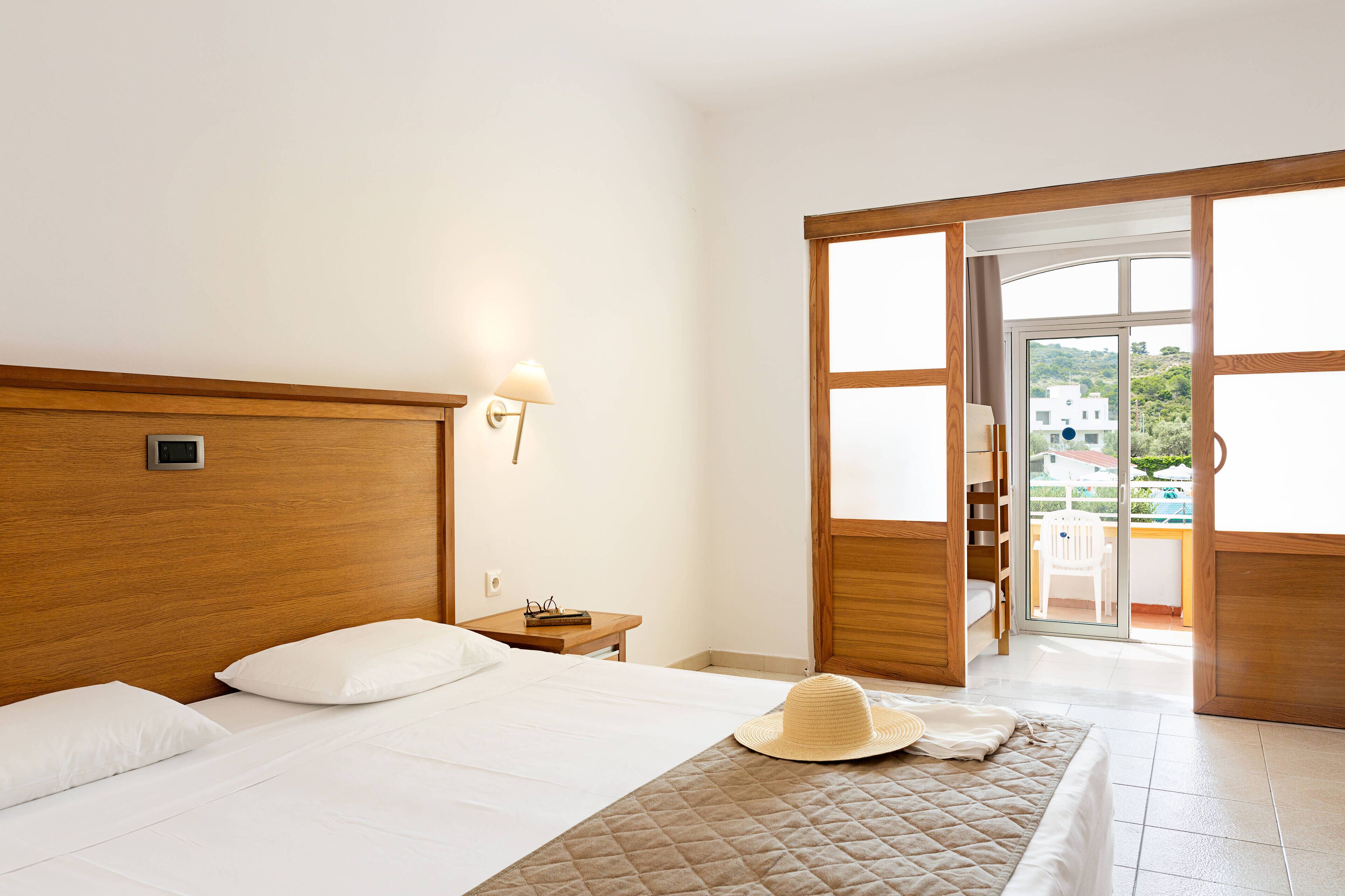 Habitación de hotel con cabecera de madera, cama con ropa blanca, mesita de noche con lámpara y vista al exterior a través de una puerta corrediza de vidrio.