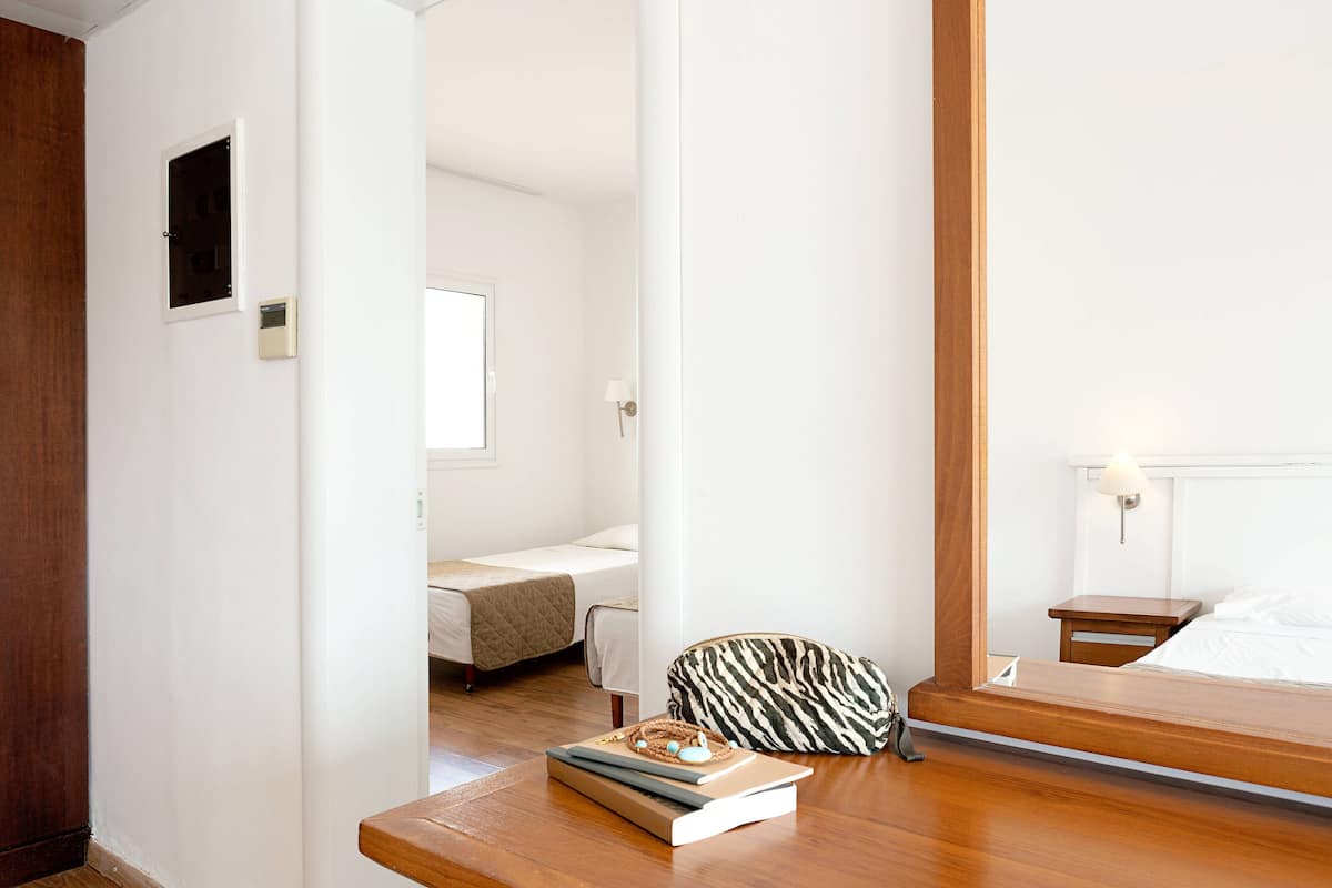 Quarto de hotel com piso de madeira, uma cama com cabeceira branca, uma mesinha de cabeceira com luminária e uma mala estampada com padrão de zebra no chão.