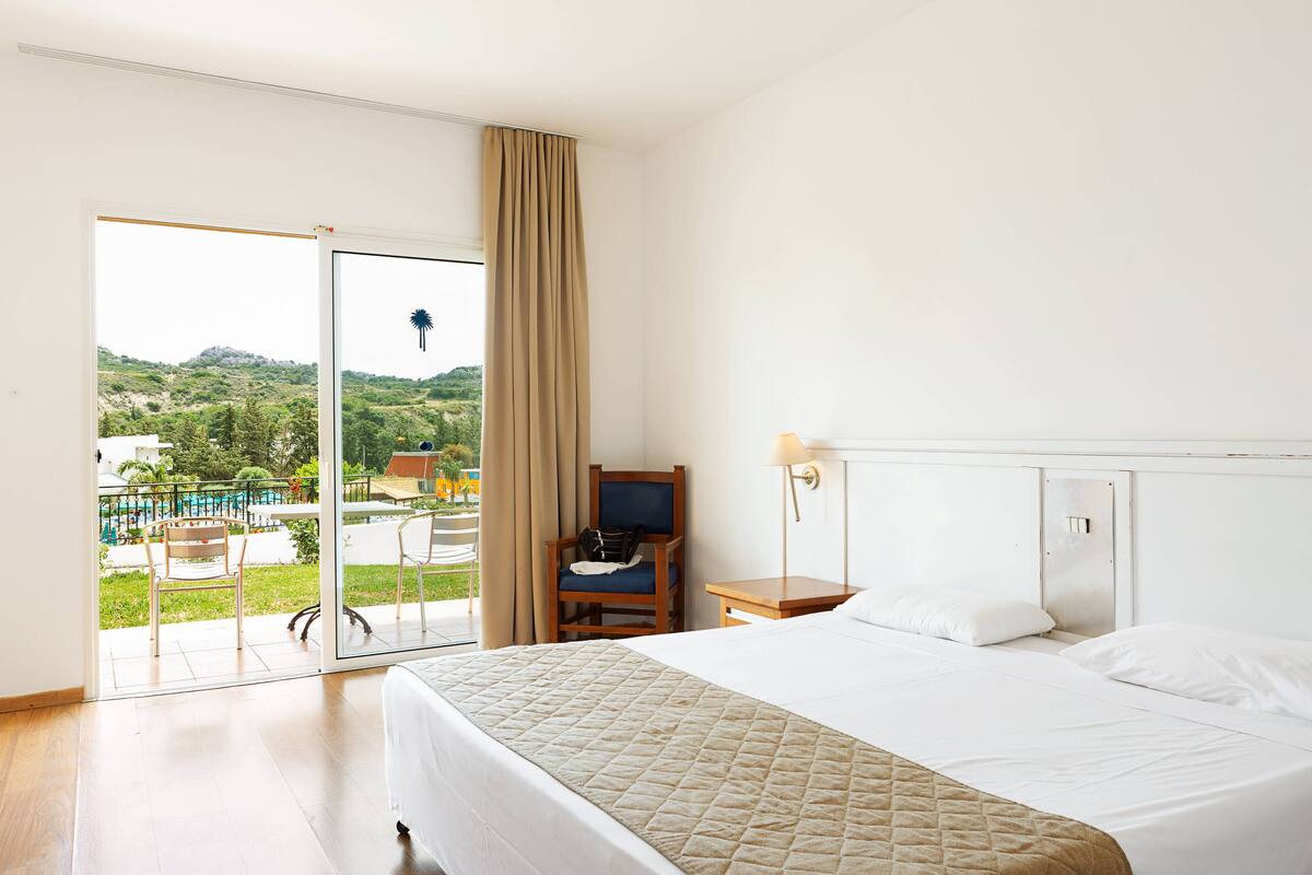 Quarto de hotel com cama, poltrona, mesa de cabeceira e vista para uma piscina e áreas verdes.