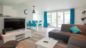 A modern living room with a flat-screen TV, a sofa, a coffee table, and a dining area with blue chairs and a table. - De Thijmse Berg (Rhenen)