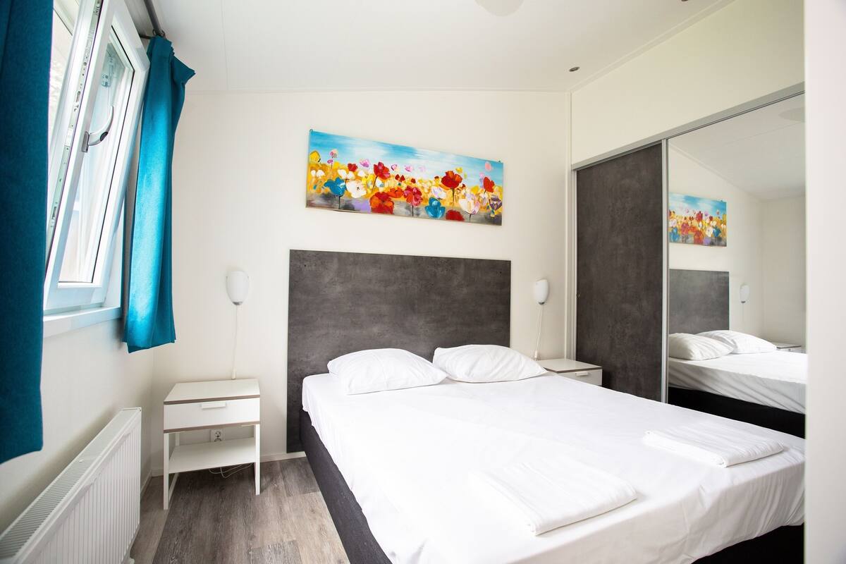 Moderni hotellihuone, jossa on suuri sänky, yöpöytä, seinälle ripustettu taulu ja suuri peili.