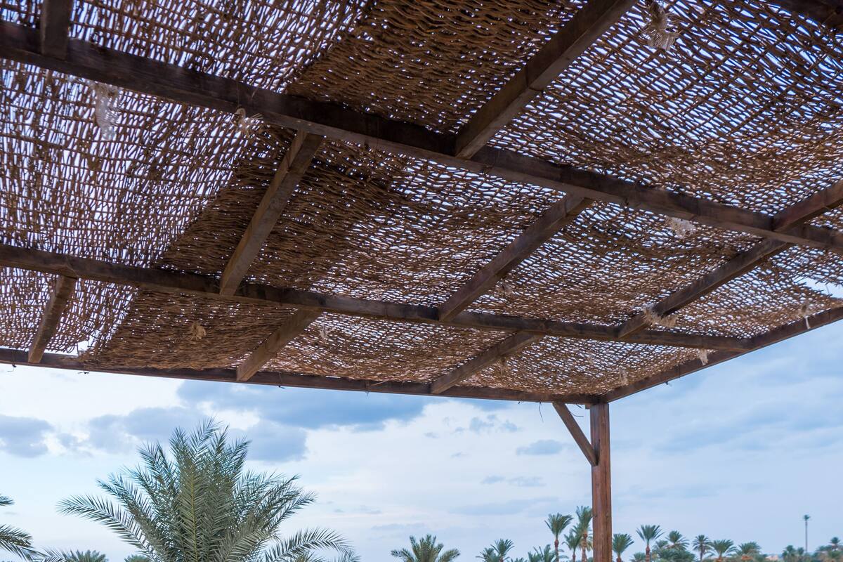 A covered outdoor seating area with a blue table and chairs, a woven ceiling, and a view of palm trees and greenery.