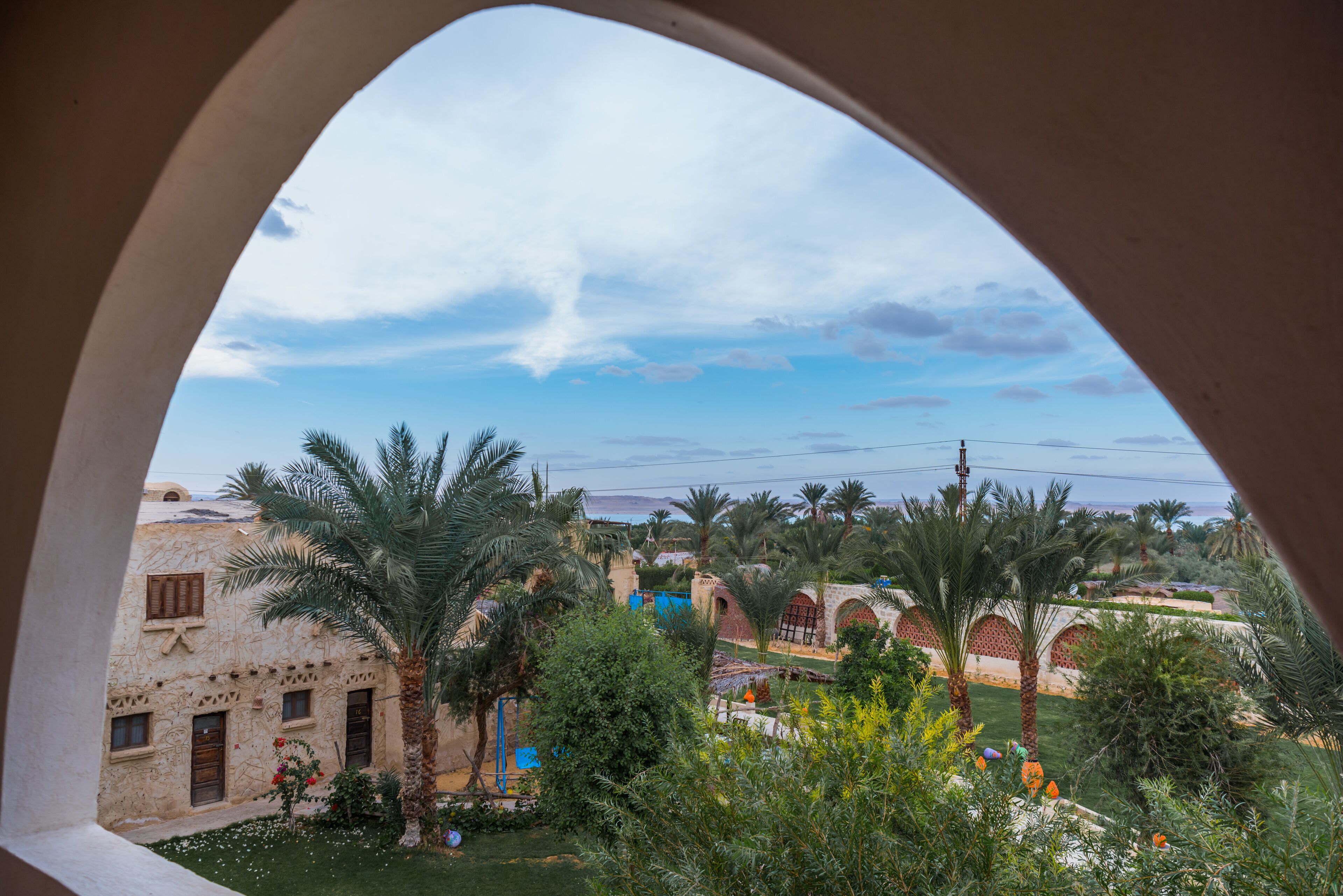 Une vue à travers une arche donnant sur une cour intérieure, avec des palmiers, un bâtiment à l’architecture traditionnelle et un ciel dégagé.