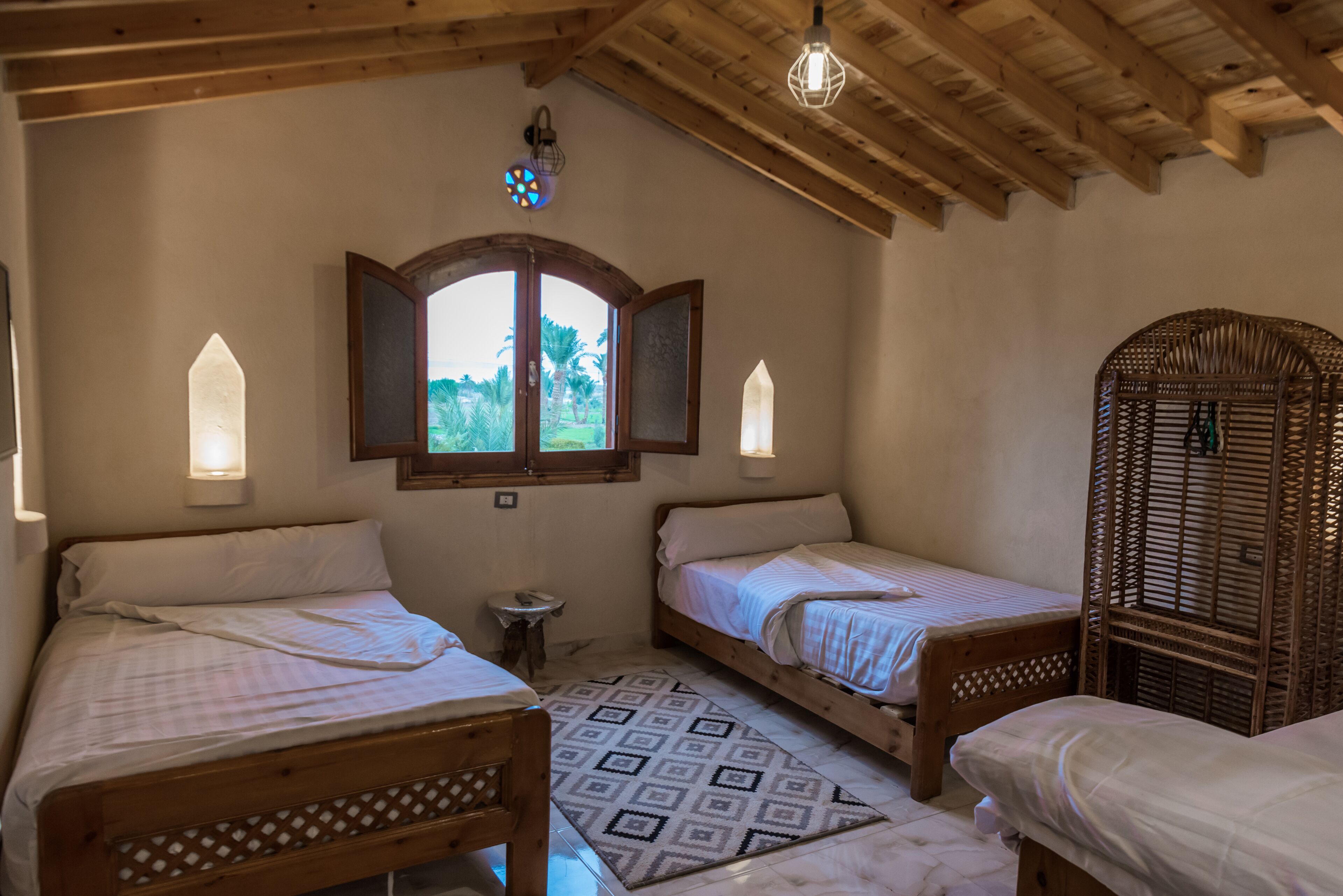 Une chambre avec deux lits simples, une petite table, un miroir décoratif et une fenêtre donnant sur un paysage verdoyant.
