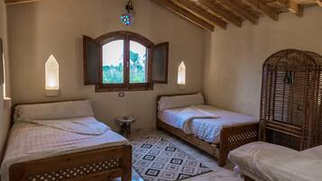 Une chambre avec deux lits simples, une petite table, un miroir décoratif et une fenêtre donnant sur un paysage verdoyant.