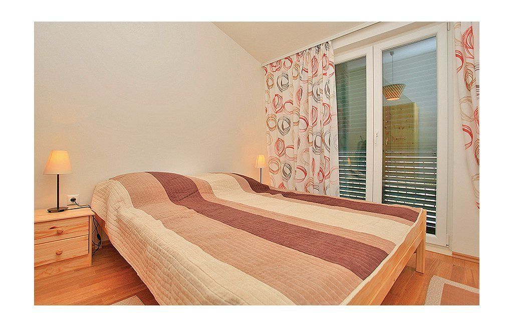 A bedroom with a bed, bedside tables, a lamp, a window with blinds, and a curtain with a pattern.