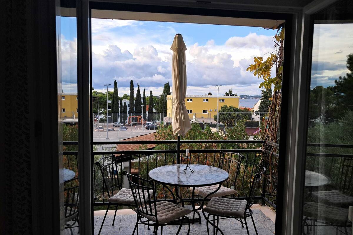 A balcony with a round table and chairs, an umbrella, and a view of a parking lot and buildings.