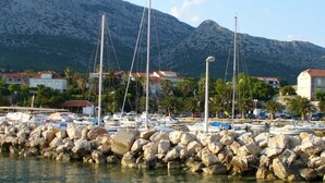 A marina with boats docked, a rocky breakwater, and houses in the background with mountains. - Apartments Trstenica (Orebic)