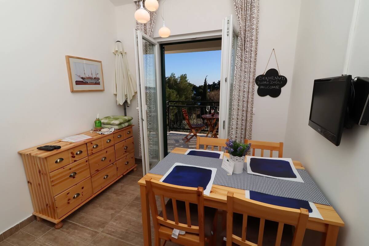 A dining area with a table set for four, wooden chairs, a small table with a plant, and a television mounted on the wall.