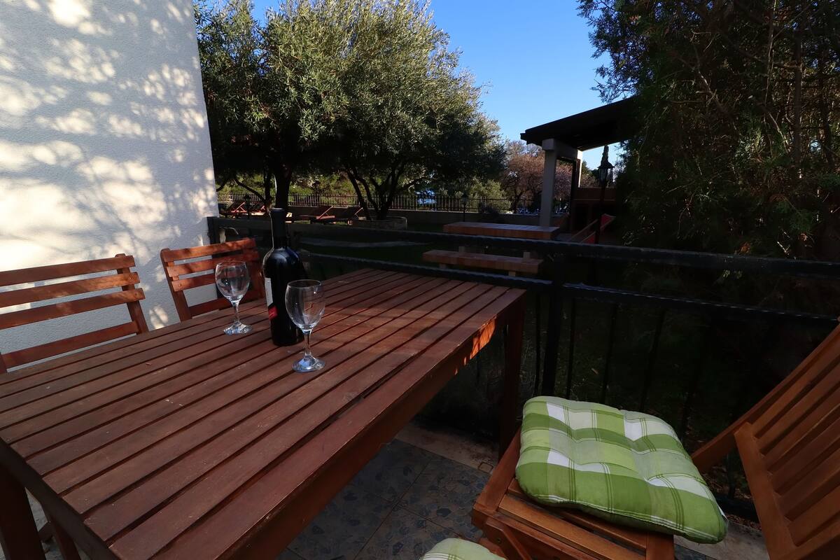 A wooden table with a wine glass and a bottle, surrounded by wooden chairs with green cushions.