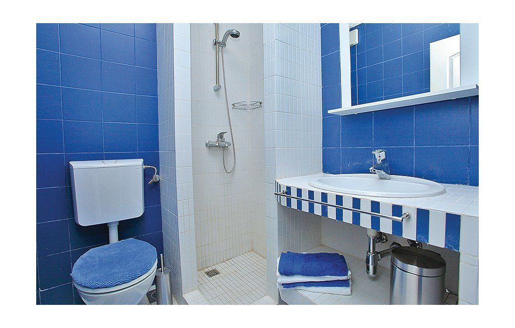 Une salle de bain avec des carreaux bleus, des toilettes blanches, une douche, un lavabo avec une serviette à rayures et une poubelle.