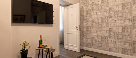 A hallway with a flat-screen TV, a small table with a bottle and glasses, a potted plant, and framed pictures on a coffee table.