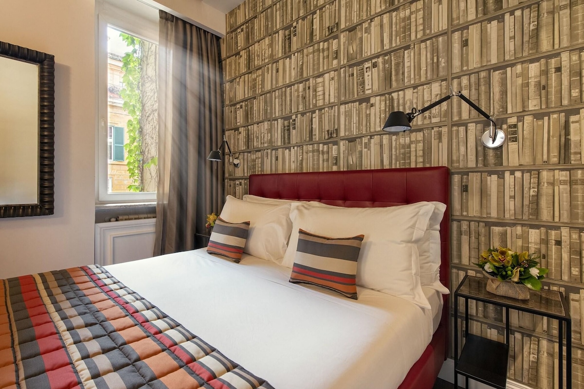 Un lit bien fait, avec une literie à rayures, une tête de lit rembourrée en tissu rouge et une table de chevet sur laquelle se trouvent une lampe et une petite plante.