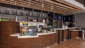 Un espace bar moderne avec un comptoir en bois, des étagères remplies de bouteilles, une machine à café et un tableau affichant le menu.