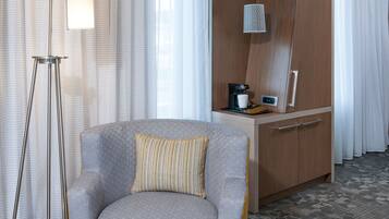 Una habitación de hotel moderna con un sofá gris, un pequeño reposapiés, una lámpara de pie, un escritorio con cafetera y un espejo grande.