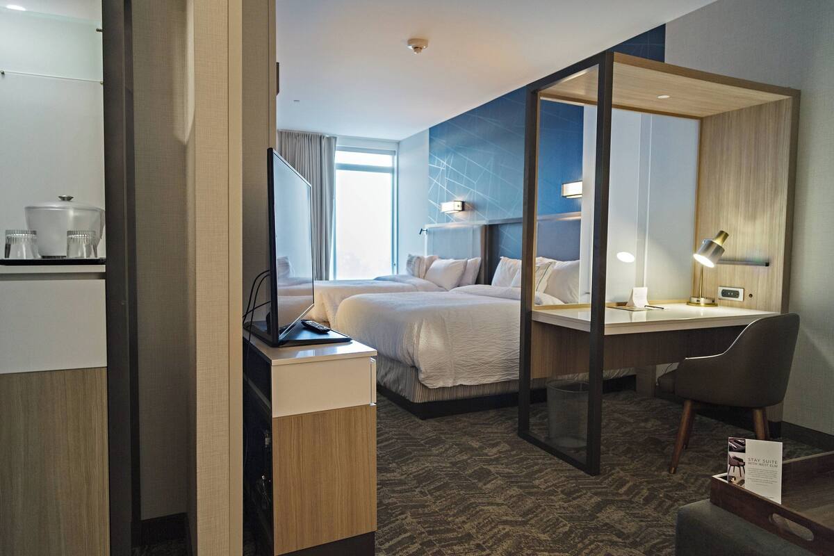 A hotel room with a bed, a desk, a chair, a mirror, and a small table with a sign.