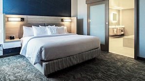 Quarto de hotel com uma cama grande, uma mesa de cabeceira, uma luminária fixada na parede e uma porta deslizante para um banheiro.