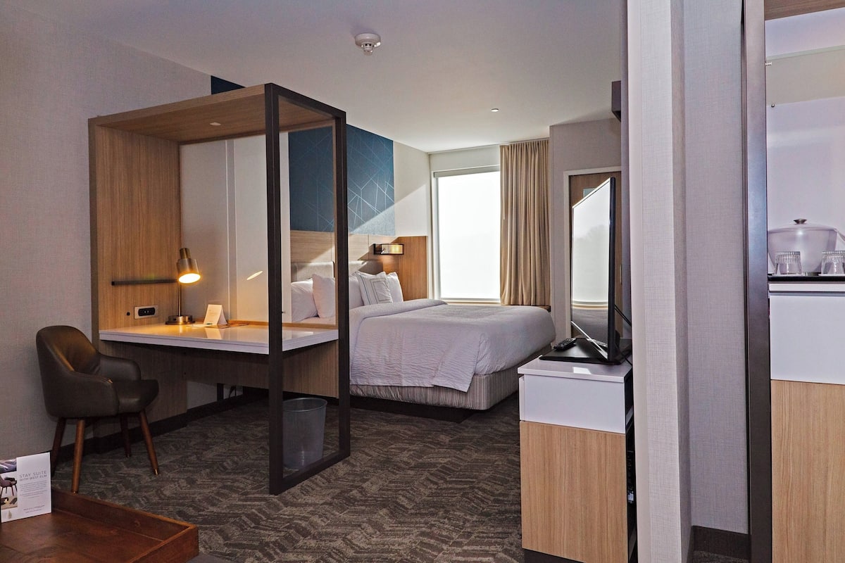 A hotel room with a bed, a desk, a chair, a mirror, and a small kitchenette.