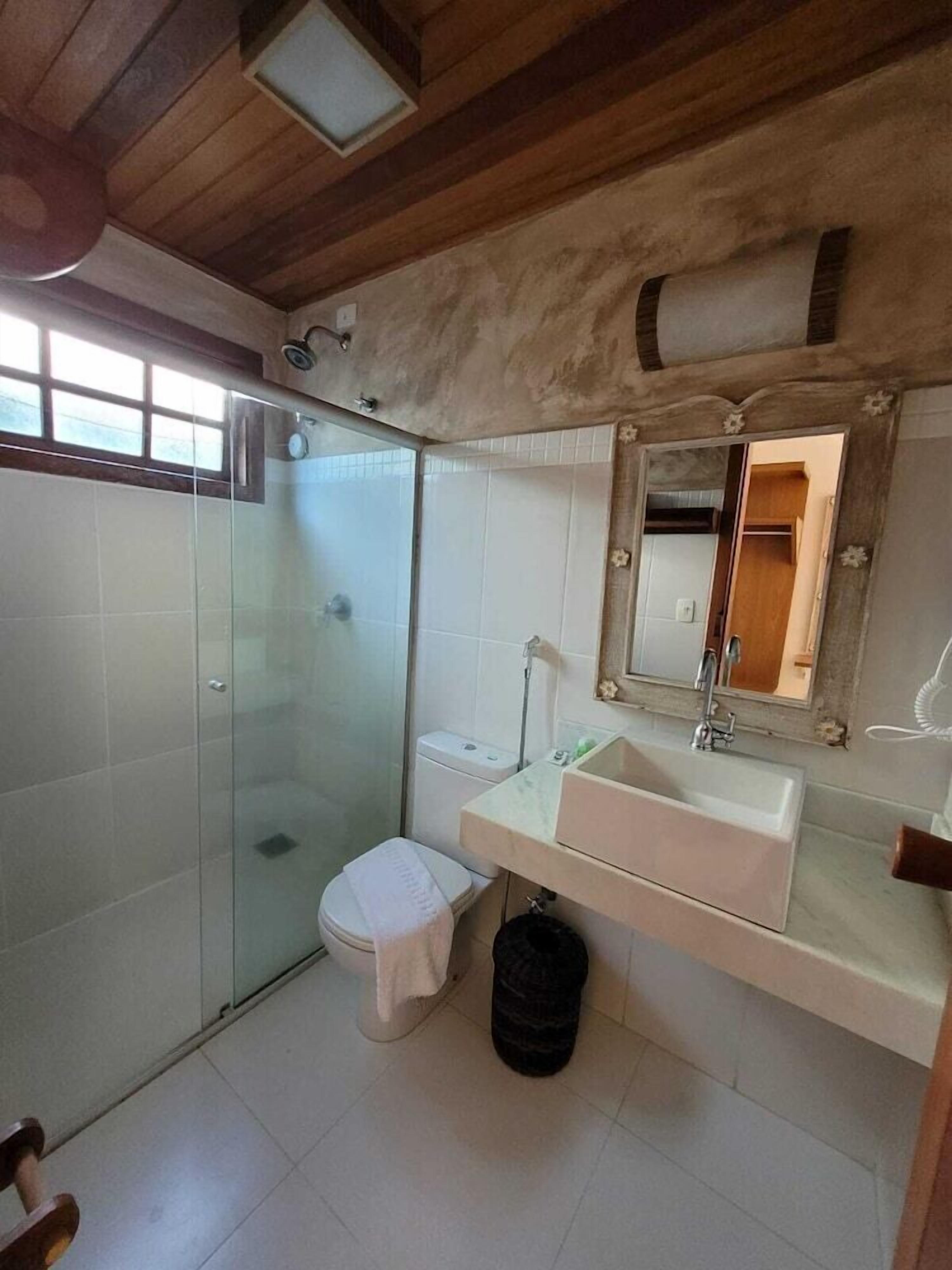 A bathroom with a glass shower enclosure, a toilet, a sink with a mirror, and a wooden ceiling.