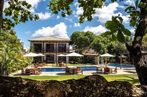 A poolside area with lounge chairs and umbrellas, a building with a tiled roof, and lush greenery. - Aquarela Praia Hotel (Porto Seguro)