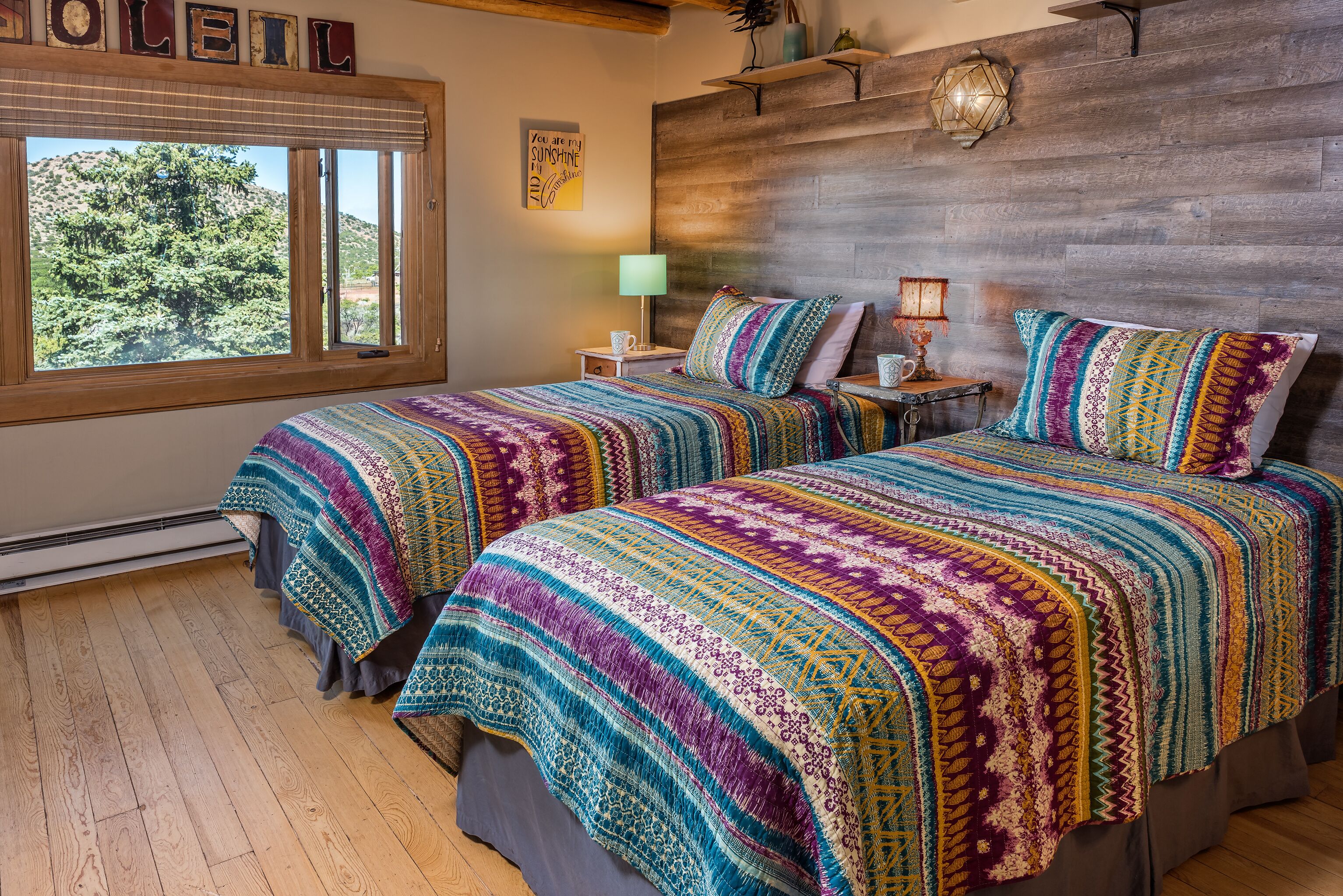 A room with two beds, a large window, a wooden headboard, and a wall-mounted lamp.