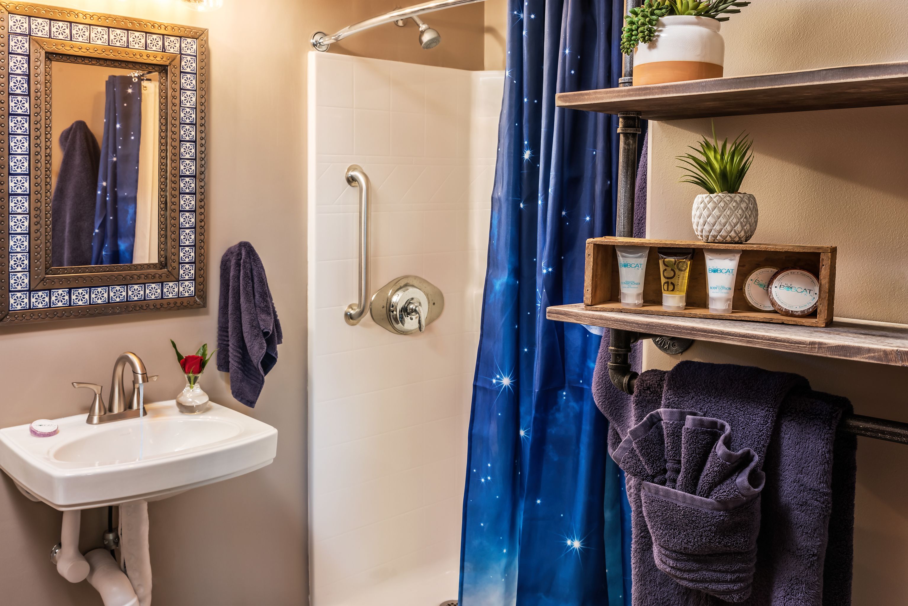 Un baño con lavamanos, espejo, ducha y repisas con artículos de aseo.