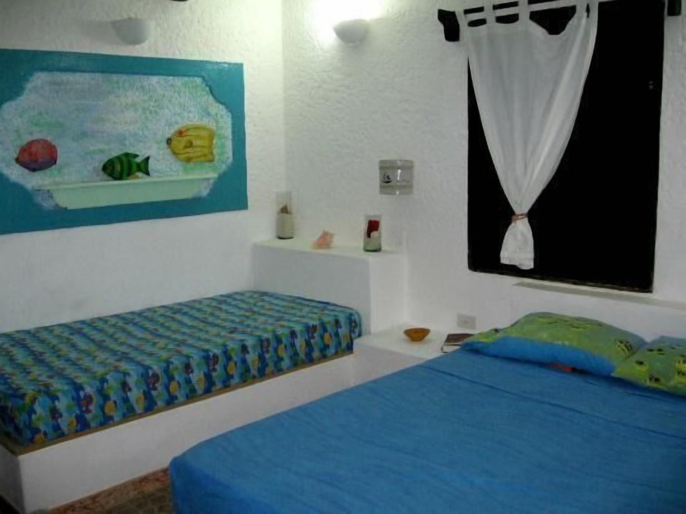 Une chambre avec deux lits, une peinture murale représentant des poissons, une petite étagère avec des articles de toilette et une fenêtre avec des rideaux blancs.