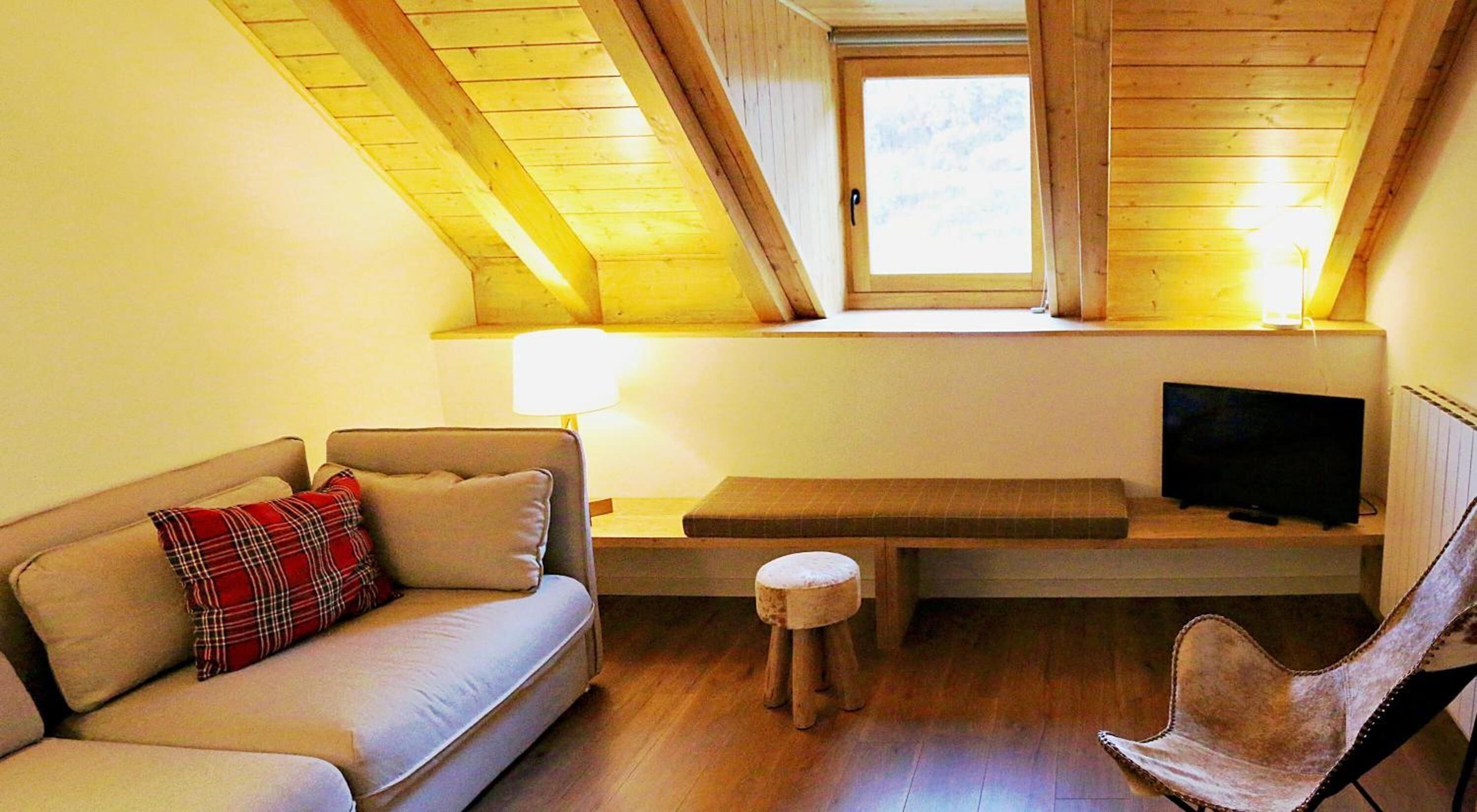 Una sala de estar acogedora con un sofá, un taburete pequeño, un banco, un televisor sobre un mueble bajo y una ventana con vigas de madera.