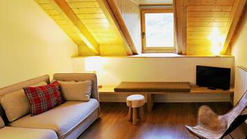Una sala de estar acogedora con un sofá, un taburete pequeño, un banco, un televisor sobre un mueble bajo y una ventana con vigas de madera.