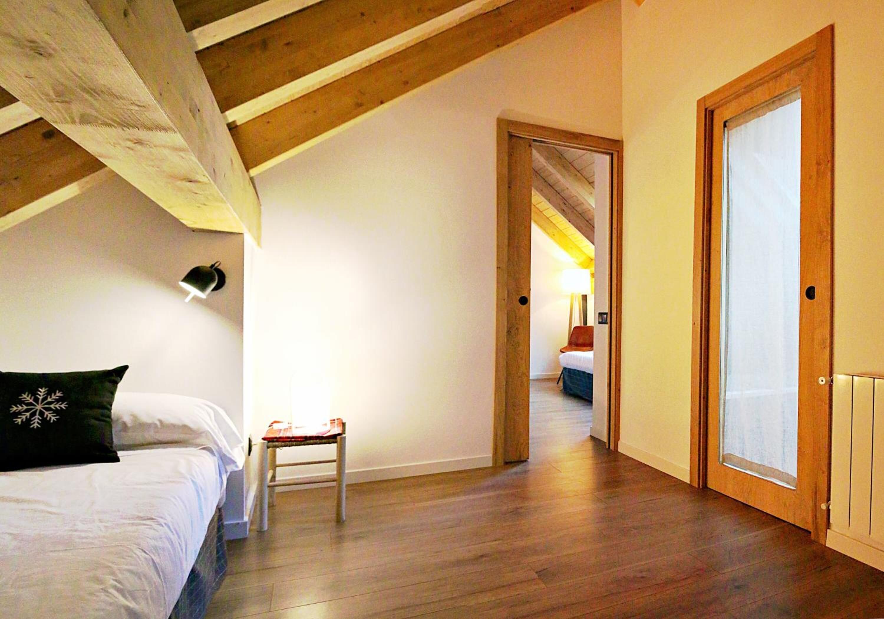 A bedroom with a wooden ceiling, a bed with white bedding, a small table, and a door leading to another room.