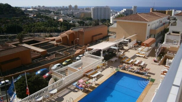 Un área de piscina en la azotea con sillones de descanso y una piscina, rodeada de edificios y vistas a la ciudad y al mar.