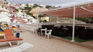 Una terraza al aire libre con sillas y mesas de plástico blanco, un área de descanso cubierta y banderas.
