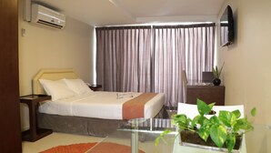 Una habitación de hotel con cama, televisor montado en la pared, aire acondicionado, ventana con cortinas y planta en maceta sobre una mesa.