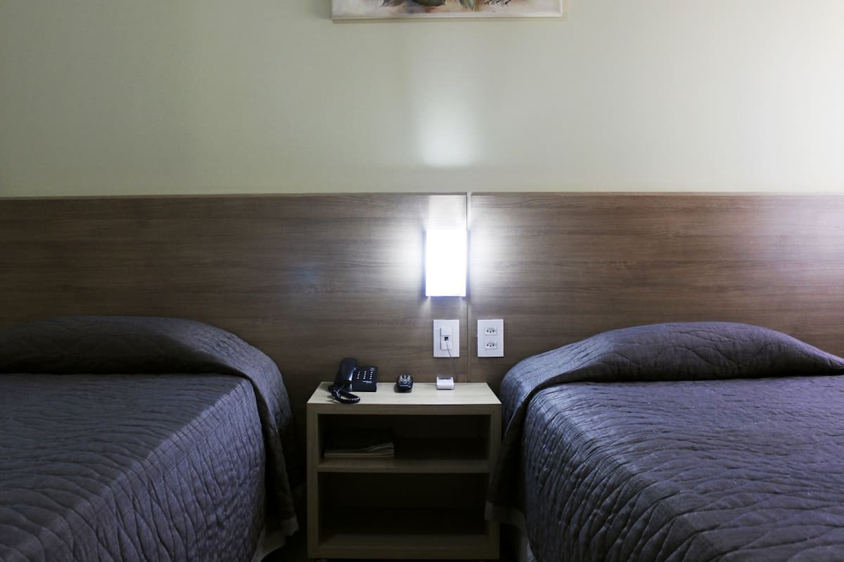 Une chambre d’hôtel avec deux lits simples, une table de chevet dotée d’un téléphone et d’une prise électrique, et une lampe fixée au mur.