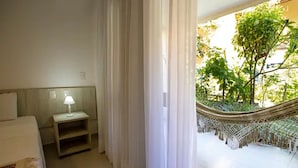 A hotel room with a bed, a nightstand with a lamp, and a view of a hammock and greenery outside. - Vivá Barra Hotel Pousada (Barra de São Miguel)