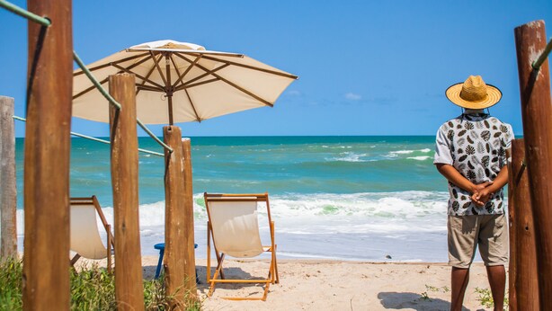 A beach scene with wooden structures, a beach umbrella, and a person standing by the ocean. - Vivá Barra Hotel Pousada (Barra de São Miguel)